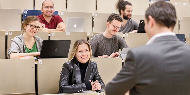 Studierende sitzen im Hörsaal auf gestaffelten Sitzreihen und hören einem Vortrag zu. Einige haben Laptops vor sich, eine Frau in der vorderen Reihe lächelt dem Vortragenden zu.