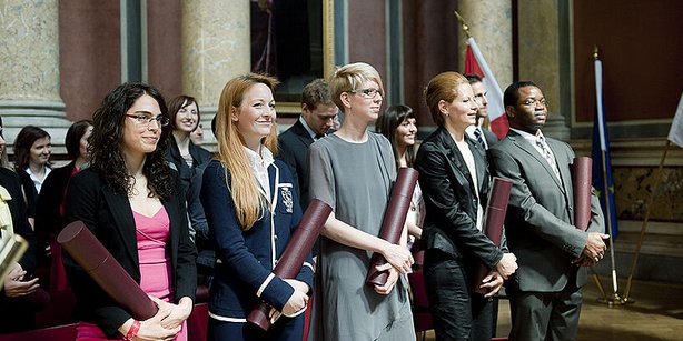Eine Gruppe von Absolvent*innen steht nebeneinander in einem prunkvollen Saal und hält gerollte Urkunden in den Händen.