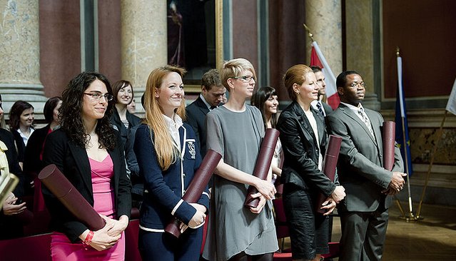 Eine Gruppe von Absolvent*innen steht nebeneinander in einem prunkvollen Saal und hält gerollte Urkunden in den Händen.