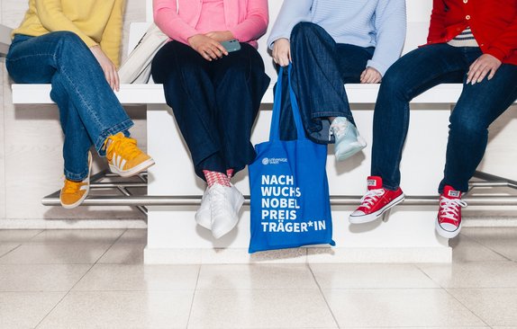 Vier sitzende Personen, nur Beine sichtbar, eine Person hält blaue Tasche mit Schriftzug "Nachwuchsnobelpreisträger*in".