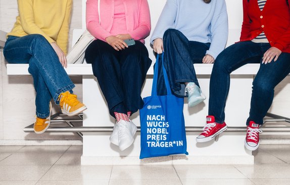 Four seated individuals, with only their legs visible; one person is holding a blue bag bearing the inscription “Nachwuchsnobelpreisträger*in”.